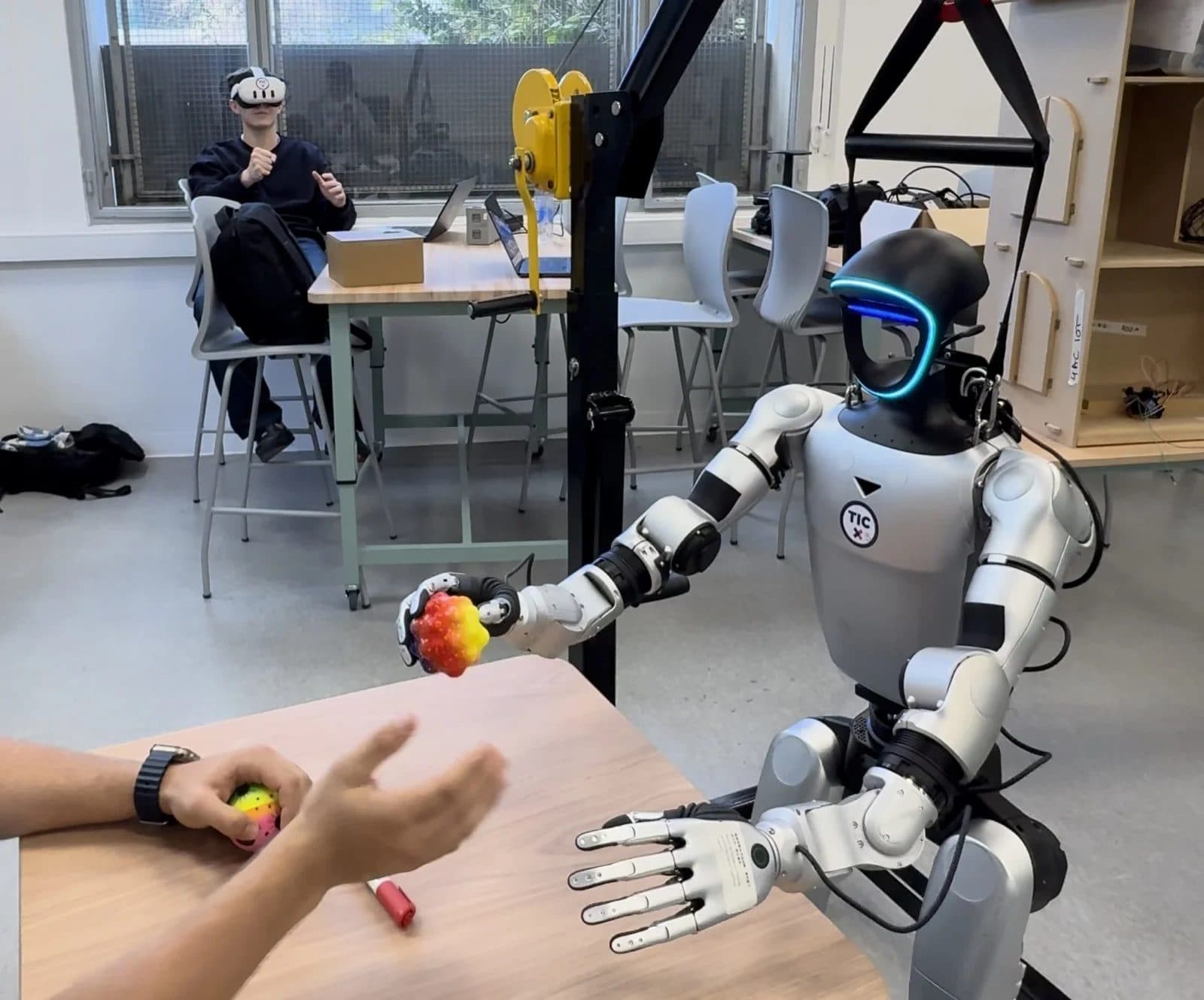 La mano del robot G1 sosteniendo una pelota mientras un operador con visor de realidad virtual la controla al fondo